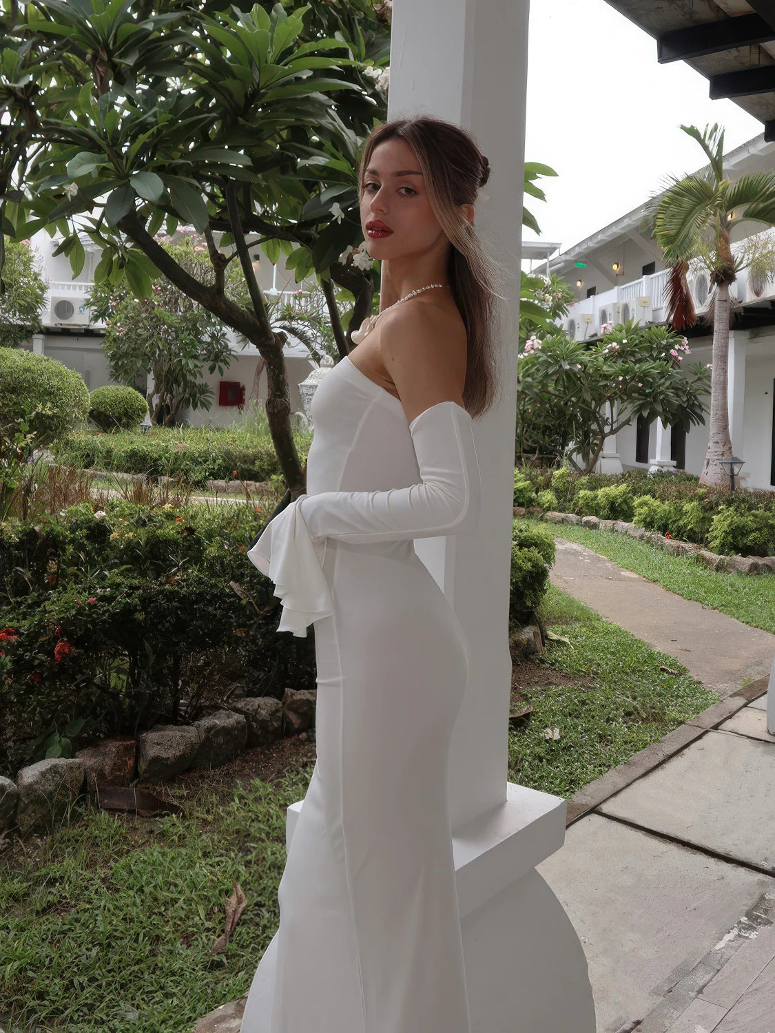 Woman in a white dress standing outdoors near a garden and building. #Color_White
