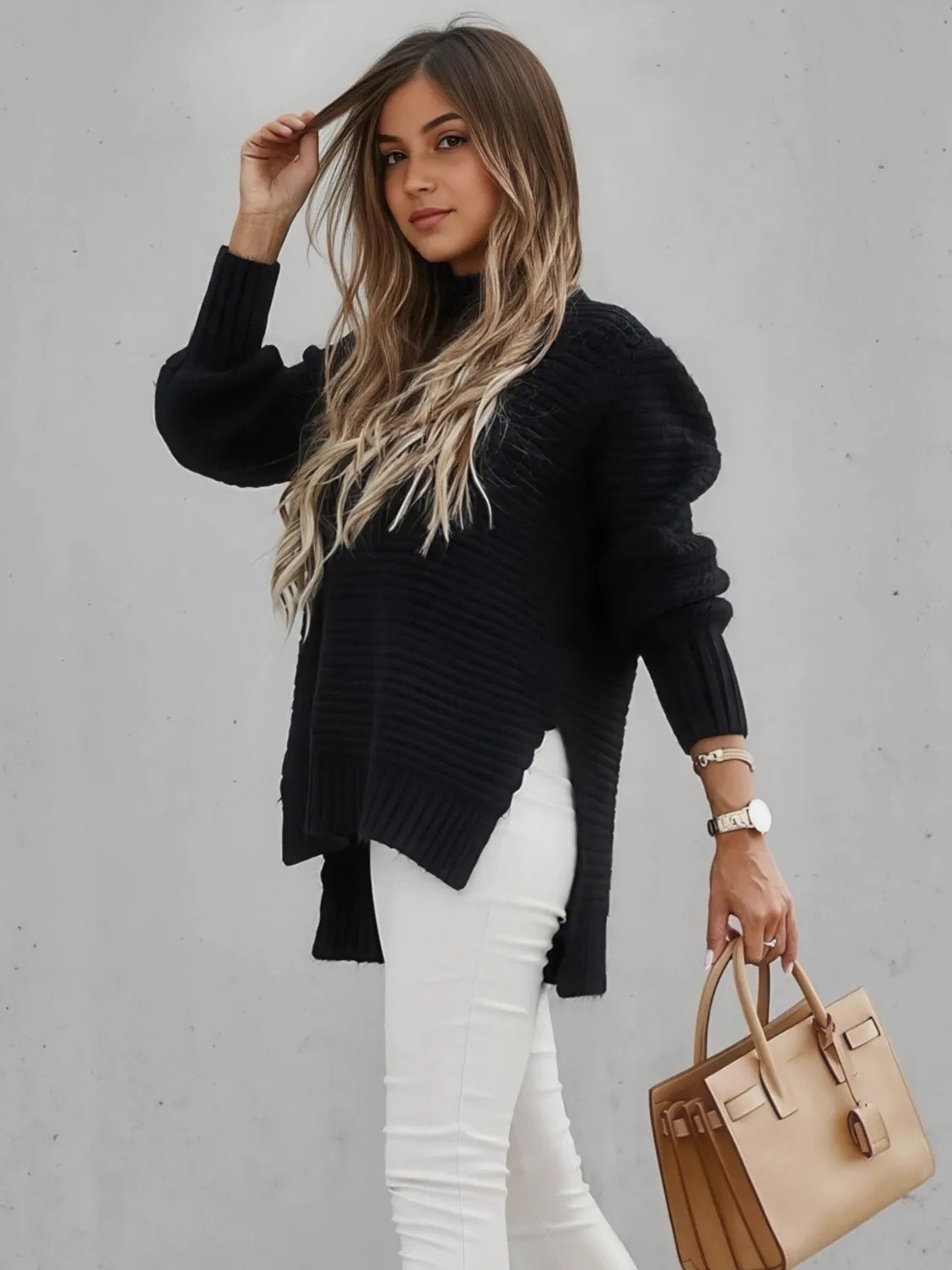 Woman wearing a black sweater and white pants, holding a beige handbag against a gray background. #Color_Black