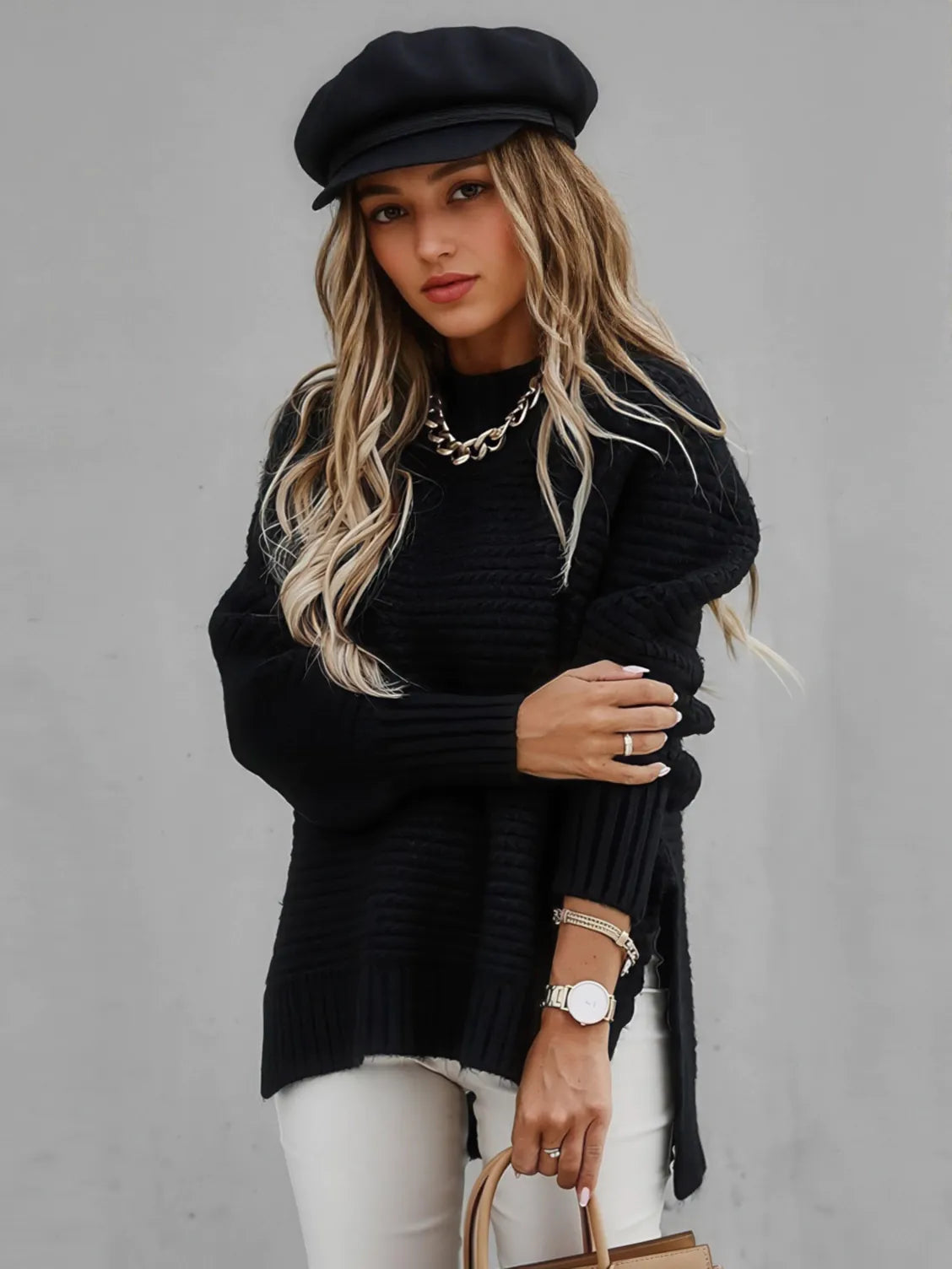 Woman wearing a black sweater, white pants, and a black cap against a gray background. #Color_Black