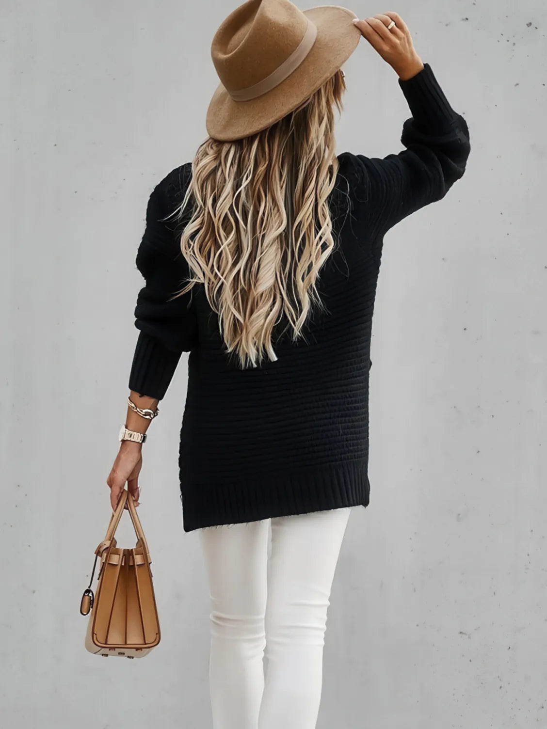 Person wearing a black sweater, white pants, and a brown hat, holding a brown handbag against a gray background. #Color_Black