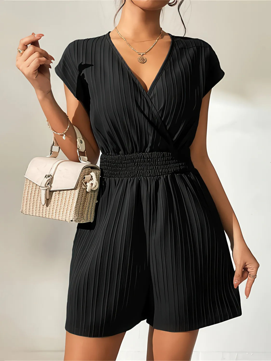 Woman wearing a black pleated romper with a beige handbag on a neutral background. #Color_Black