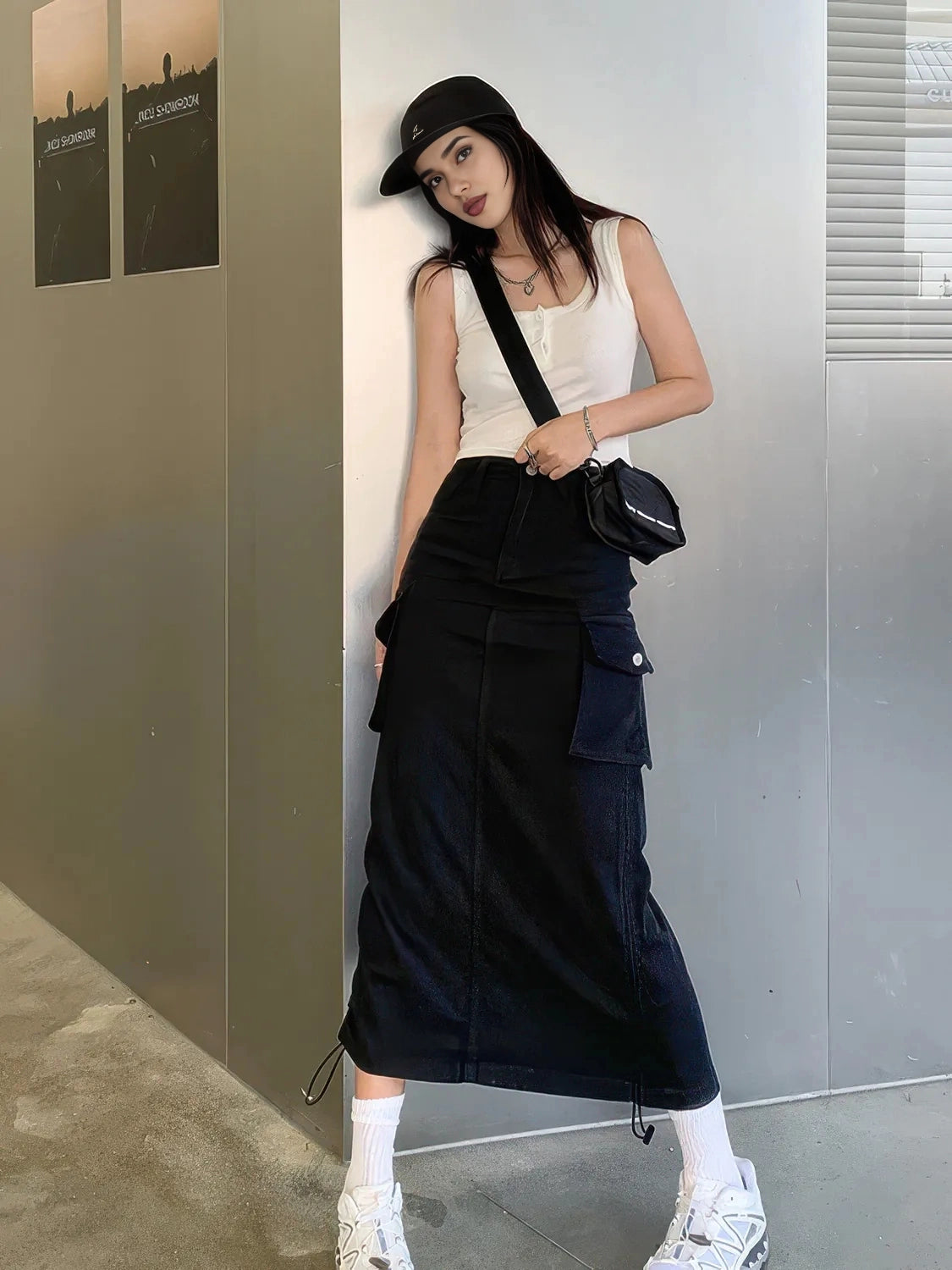 Woman in a white top and black skirt with a black cap and bag against a gray wall. #Color_Black