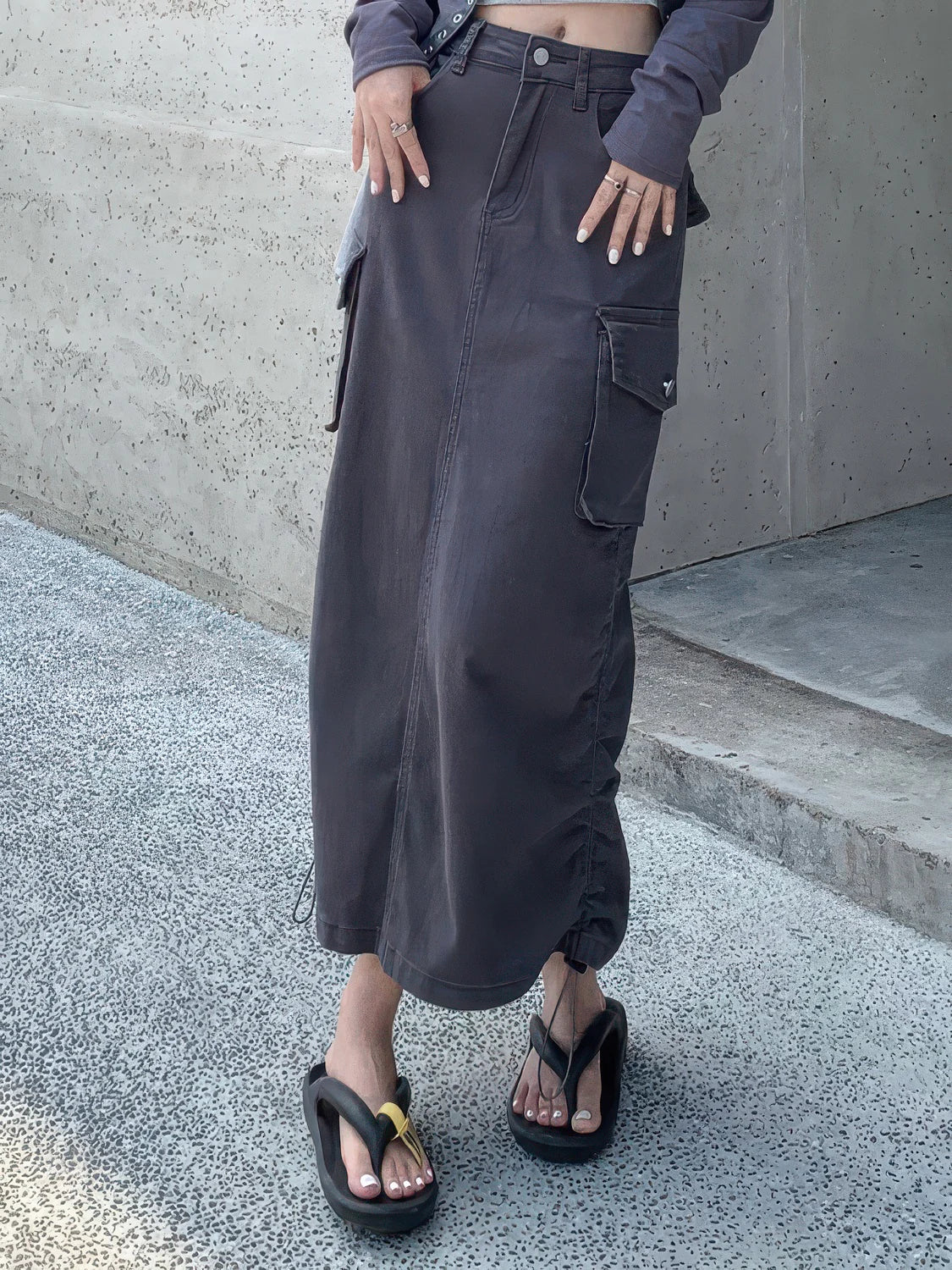 Person wearing a long dark gray skirt with a concrete wall background. #Color_Gray