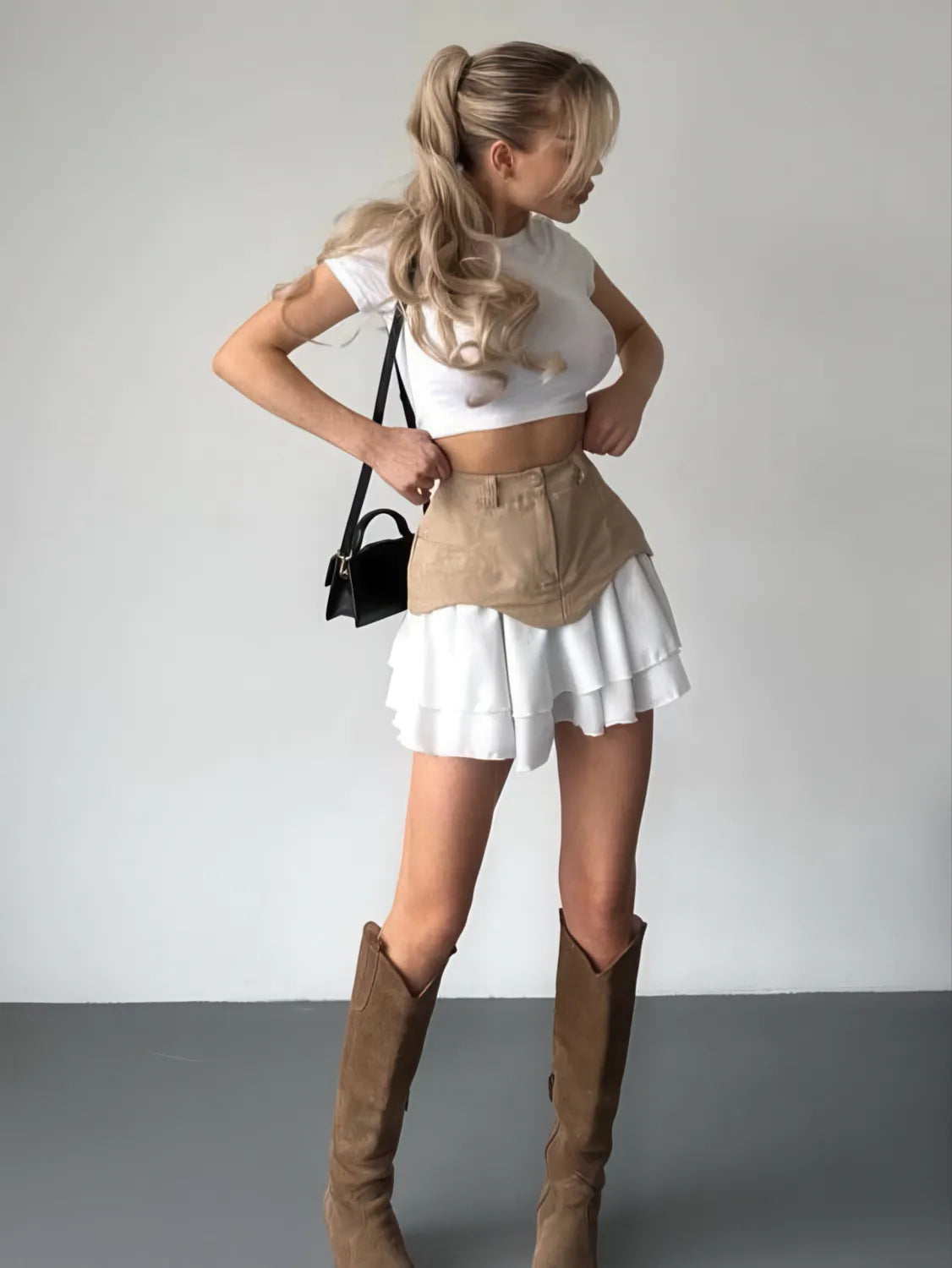 Woman wearing a white crop top, beige skirt, and brown knee-high boots against a plain background. #Color_Khaki