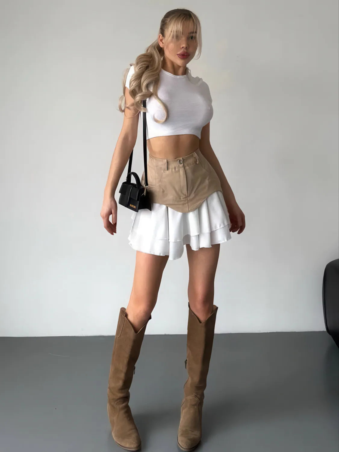 Woman wearing a white crop top, beige skirt, and brown knee-high boots against a plain background. #Color_Khaki