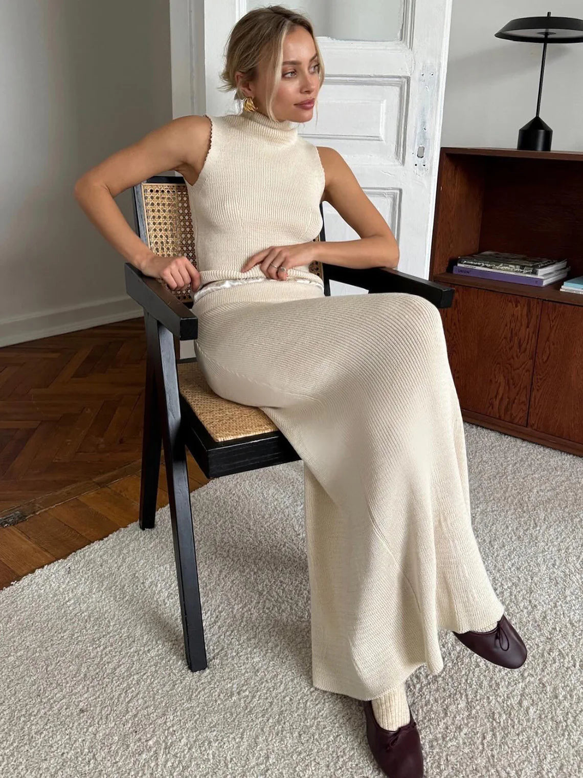 Woman sitting on a chair wearing a beige sleeveless top and long skirt in a room with wooden floor and furniture. #Color_Beige