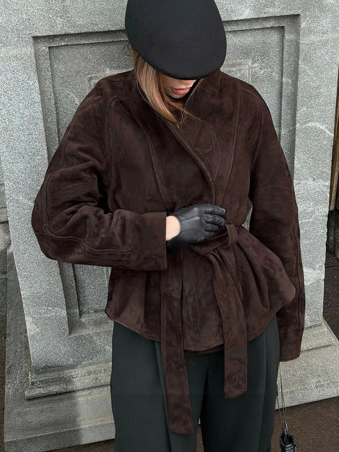 Person wearing a brown suede jacket and black gloves against a gray stone wall. #Color_Brown